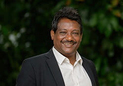 A man wearing a dark blazer and white shirt is smiling in front of a blurred green leafy background.