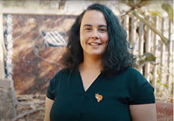 Woman with curly dark hair wearing a black shirt and fox pin, standing outdoors with a painting and wooden fence in the background.