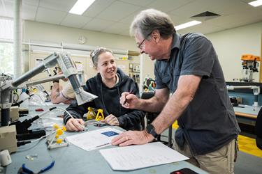 An instructor points to a circuit board diagram on a workbench while explaining it to a student in a laboratory.