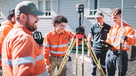 Four surveyors in high-visibility workwear gathered around a Trimble surveying instrument mounted on a tripod.