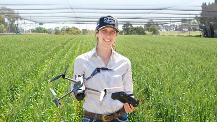 A woman in an Ariat cap standing in a wheat field, holding a drone and its remote controller.