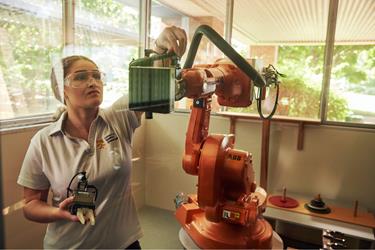 A student wearing safety goggles adjusts the end effector of an orange ABB industrial robotic arm.