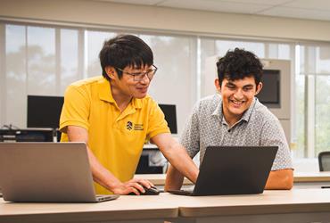 A student in a yellow polo shirt and another student looking at a laptop together at a desk.