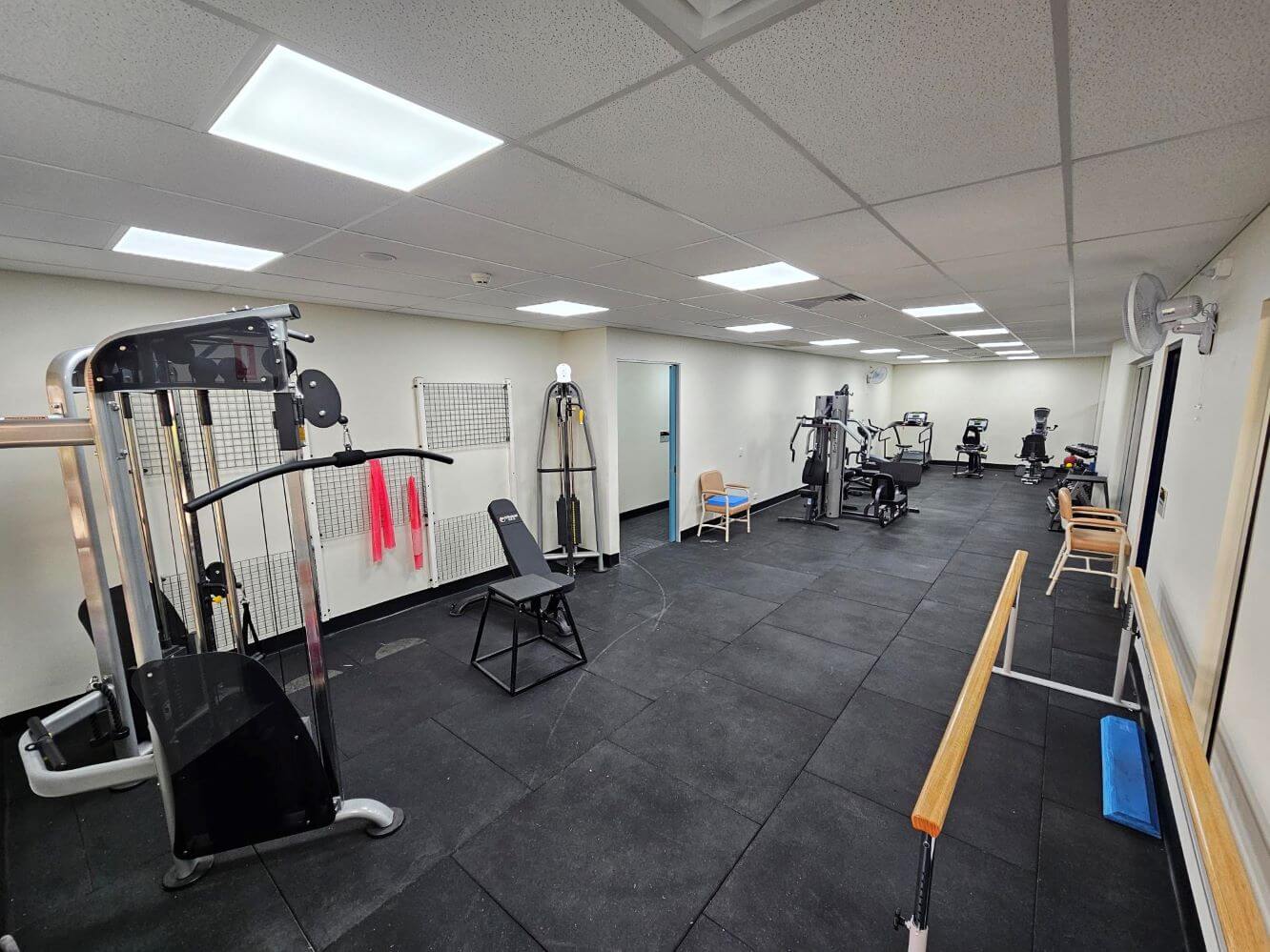 A gym room with various exercise equipment including weight machines, stationary bikes, benches, and resistance bands on a rack. The floor is covered with black mats.