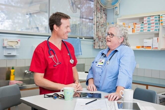 Two healthcare professionals, one in red scrubs and one in blue, are standing and talking in a medical office next to paperwork, a tablet, and mugs.