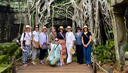 A group of nine people pose in front of large tree roots covering stone ruins, surrounded by greenery.