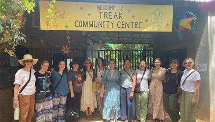 A group of thirteen people stands smiling in front of the Treak Community Centre entrance, beneath a yellow welcome banner, with plants and colorful decorations around them.