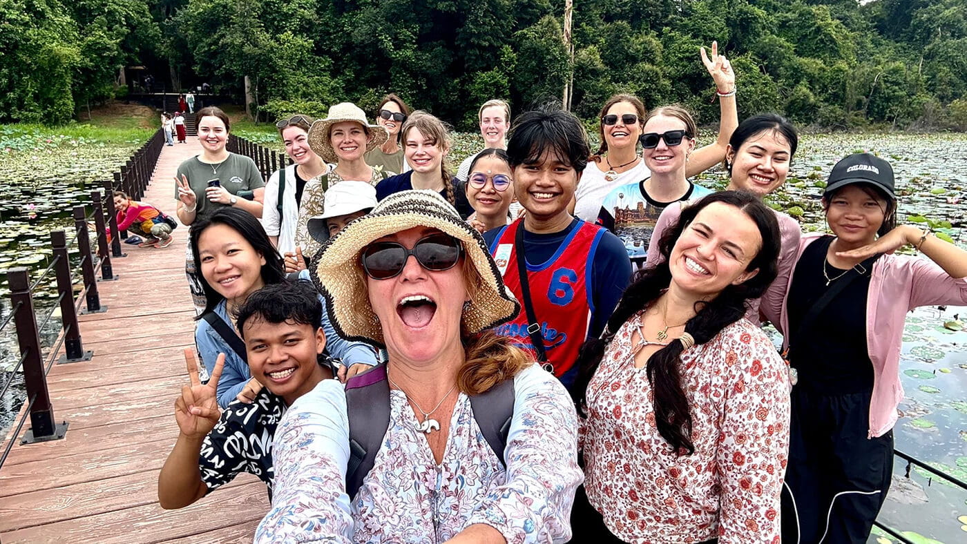 A group of people smiling and posing for a selfie on a wooden walkway over a lily pond, with dense green trees in the background.