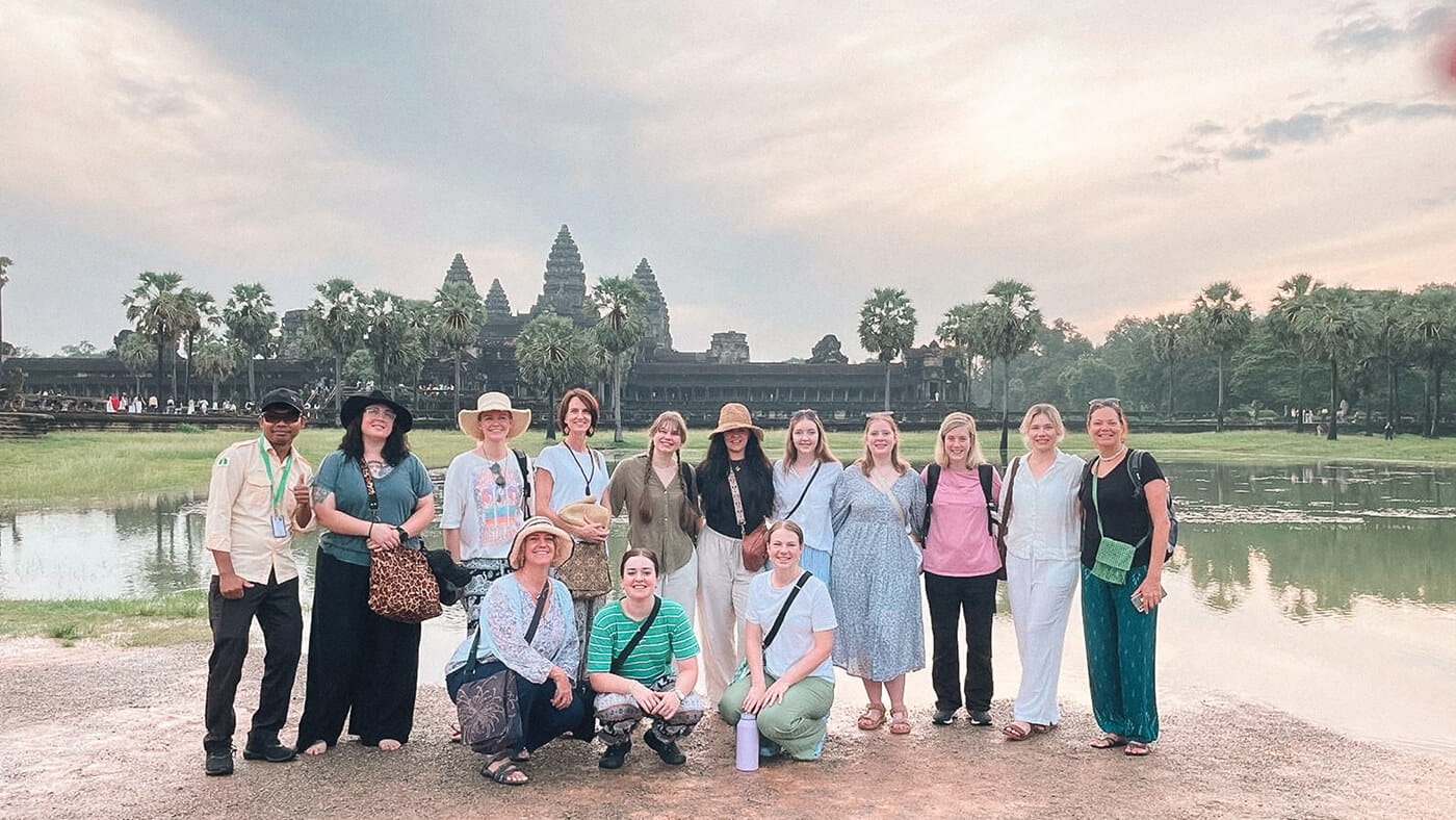 A group of people poses in front of Angkor Wat temple, with palm trees and a pond in the background under a cloudy sky.