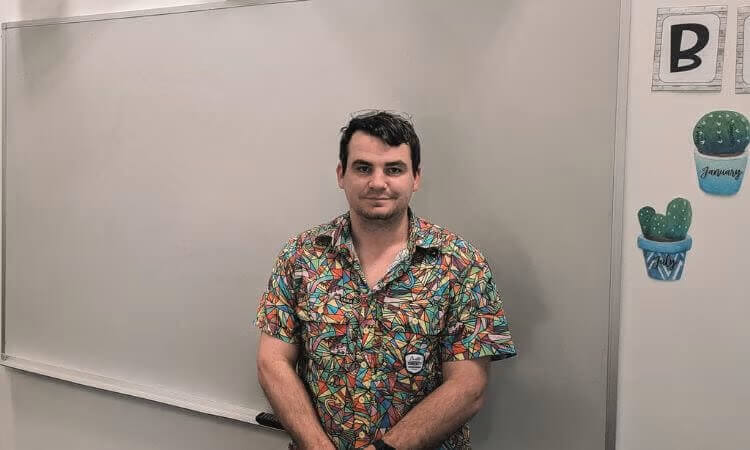 A man in a colorful shirt stands in front of a blank whiteboard, with cactus decorations and the letter B displayed on the wall to his right.