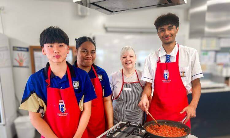 Four people in aprons stand in a kitchen. One person is stirring food in a pan on the stove. They are smiling at the camera.