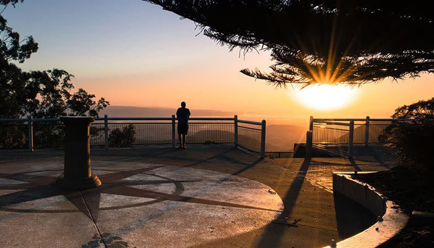 A person stands at a railing overlooking a scenic view during sunset, with trees and shadows visible in the foreground.