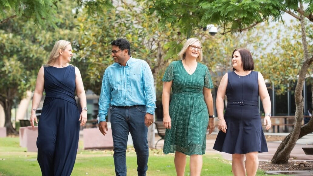 Four adults walk together outdoors on a grassy area, smiling and talking, with trees and benches in the background.