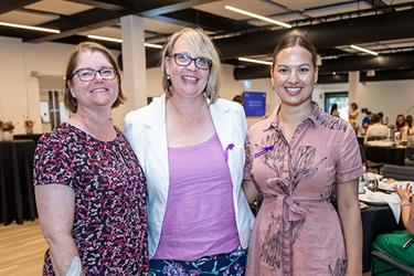 Three women smiling at a professional event, each wearing a different outfit, with one in a floral dress, one in a white blazer, and one in a pink dress. They are indoors with other people in the background.