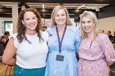 Three women standing indoors, smiling at the camera. They are each wearing purple ribbons and appear to be attending an event.