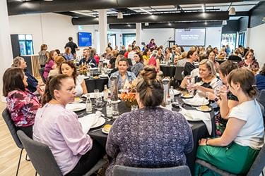 A large group of people are seated at round tables in a bright, modern conference room. They are engaged in conversation, eating, and drinking. A presentation screen is visible in the background.