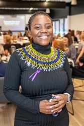 A woman in a black dress with colorful beaded neckpiece stands smiling indoors at an event, holding a folded pair of glasses with people seated at tables in the background.