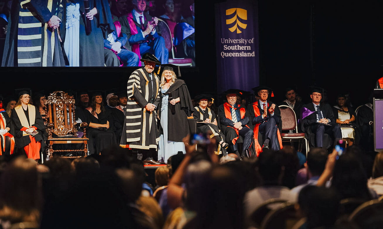 A graduation ceremony at the University of Southern Queensland with graduates and faculty on stage, and an audience watching and taking photos.