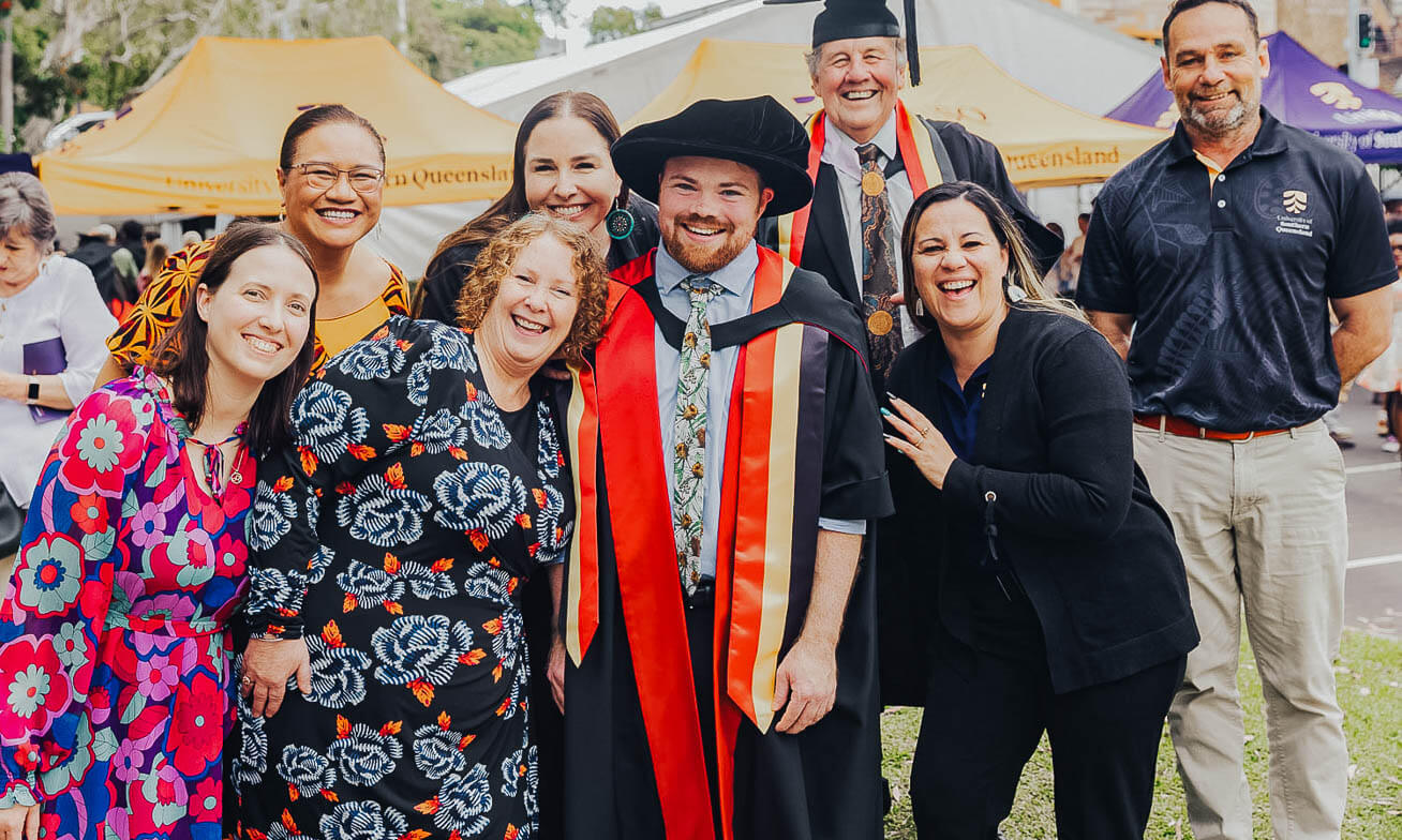 A group of people smile and pose outdoors at a graduation event, with one person in academic regalia standing in the center.
