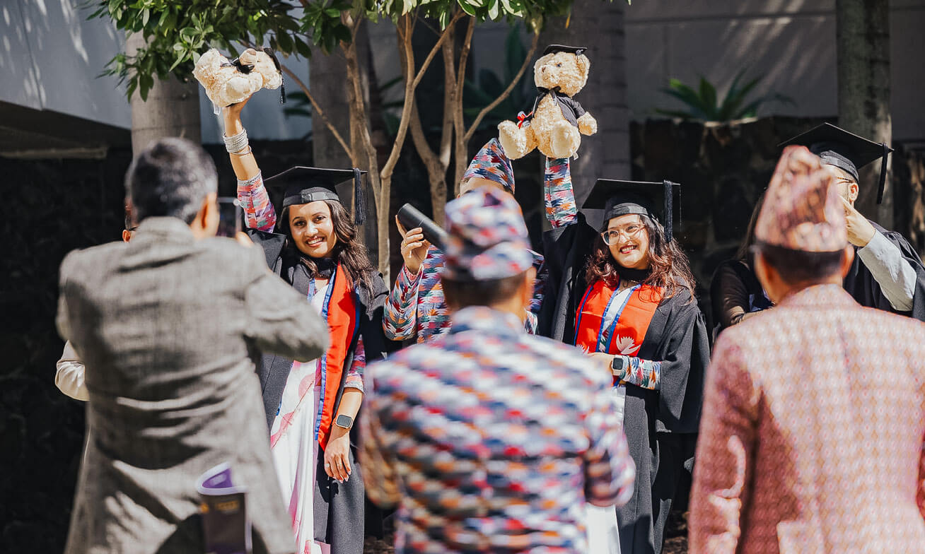 Two graduates in caps and gowns smile and hold up stuffed bears while people in patterned clothing take photos of them outdoors.