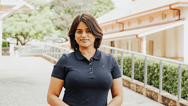 A person with short dark hair, wearing a navy blue polo shirt, stands outdoors near a walkway with greenery and buildings in the background.