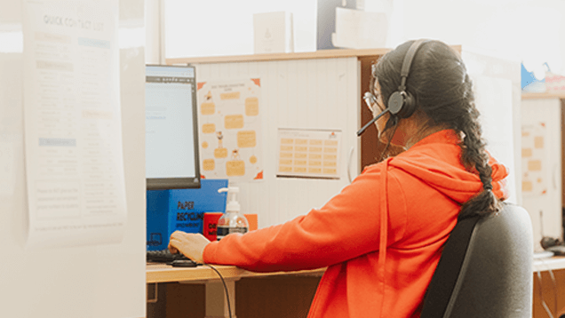 Person wearing a headset and red hoodie sits at a computer workstation in an office, with a hand sanitizer bottle on the desk.