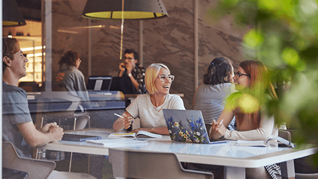 Three people sit around a table indoors, talking and smiling, with notebooks and a laptop in front of them; other people are seated in the background.