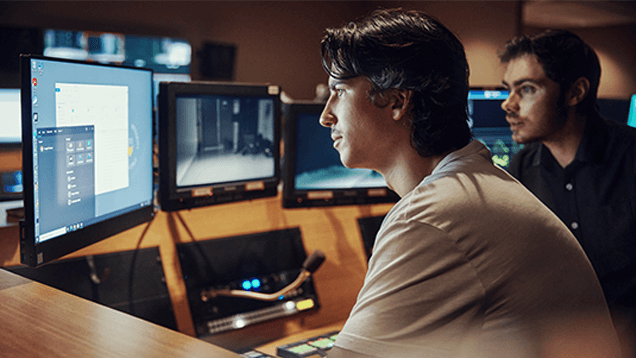 Two people sit at a control desk with multiple monitors displaying various screens in a dimly lit room.