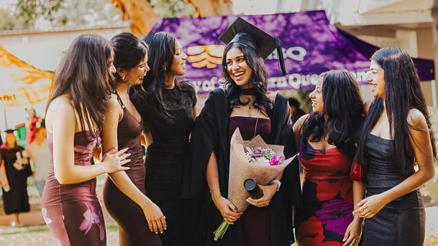 A group of young women stand together outdoors, smiling. One wears a graduation cap and gown, holding a bouquet and diploma, while the others wear dresses.
