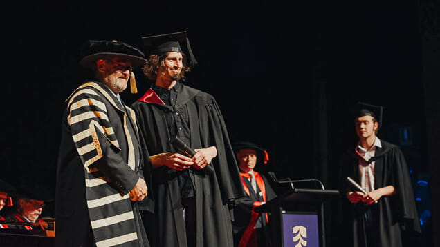 A graduate in a cap and gown stands on stage holding a diploma beside an official in ceremonial robes during a graduation ceremony.
