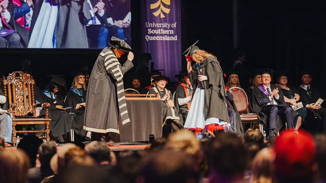 A graduate bows to an official on stage during a graduation ceremony at the University of Southern Queensland, with an audience and faculty members seated and clapping.