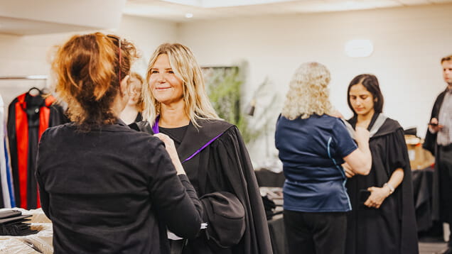 Several people are preparing for a graduation ceremony, adjusting their gowns and talking in a brightly lit indoor space.