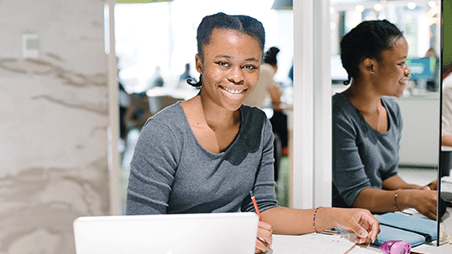 A person sits at a desk with a laptop, notebook, and pen, smiling at the camera; their reflection is visible in a glass wall beside them.