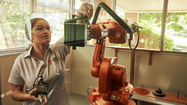 A woman in safety glasses operates an industrial robotic arm inside a workshop with large windows and natural light.