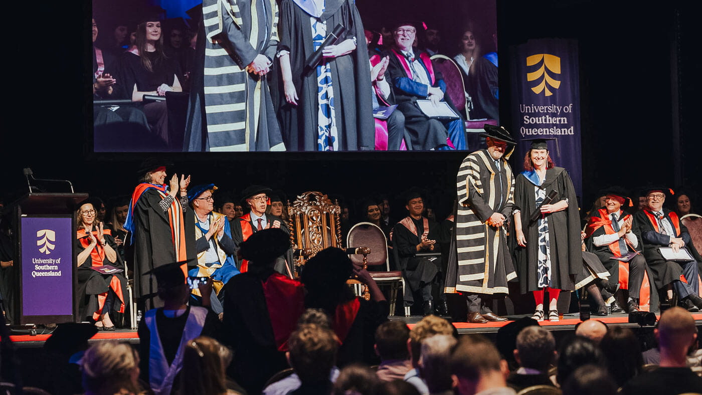 A graduate receives a diploma on stage during a University of Southern Queensland graduation ceremony, with officials and audience members present.