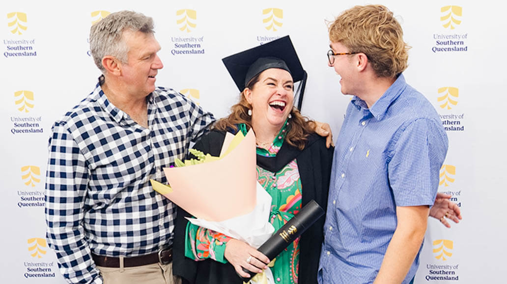 Three people stand together smiling in front of a University of Southern Queensland backdrop; one in graduation attire holds a bouquet and a diploma.