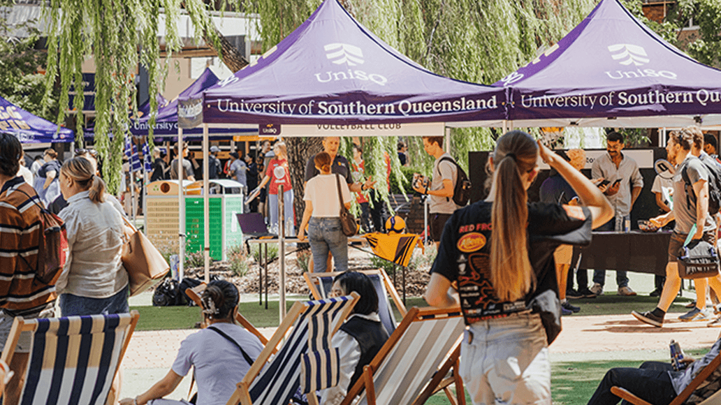People gather outdoors near University of Southern Queensland tents, with chairs, informational tables, and various activities visible under sunny weather.