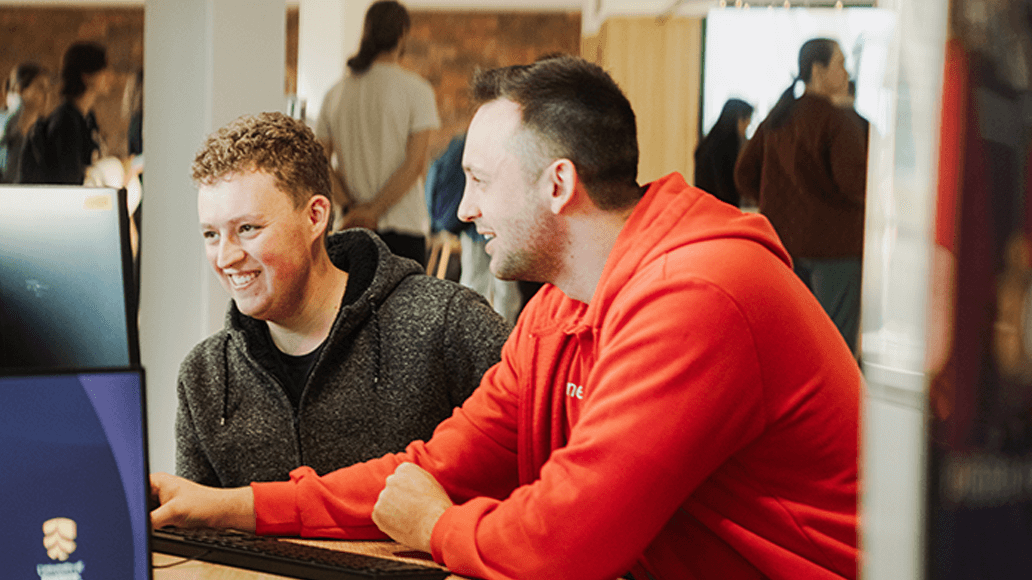 Two men sitting side by side at a computer desk, smiling and looking at the monitor in a busy indoor setting.