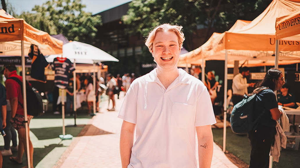 A person in a white shirt stands smiling outdoors at an event with several people and yellow tents in the background.