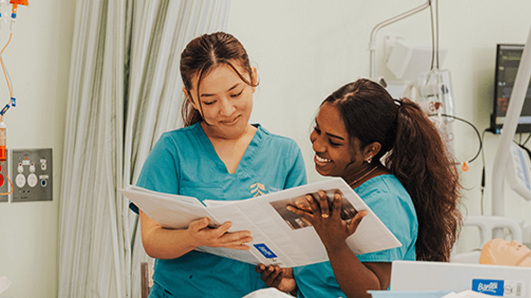 Two female UniSQ nursing students in teal scrubs stand together in a medical setting, reading and discussing a binder while smiling.