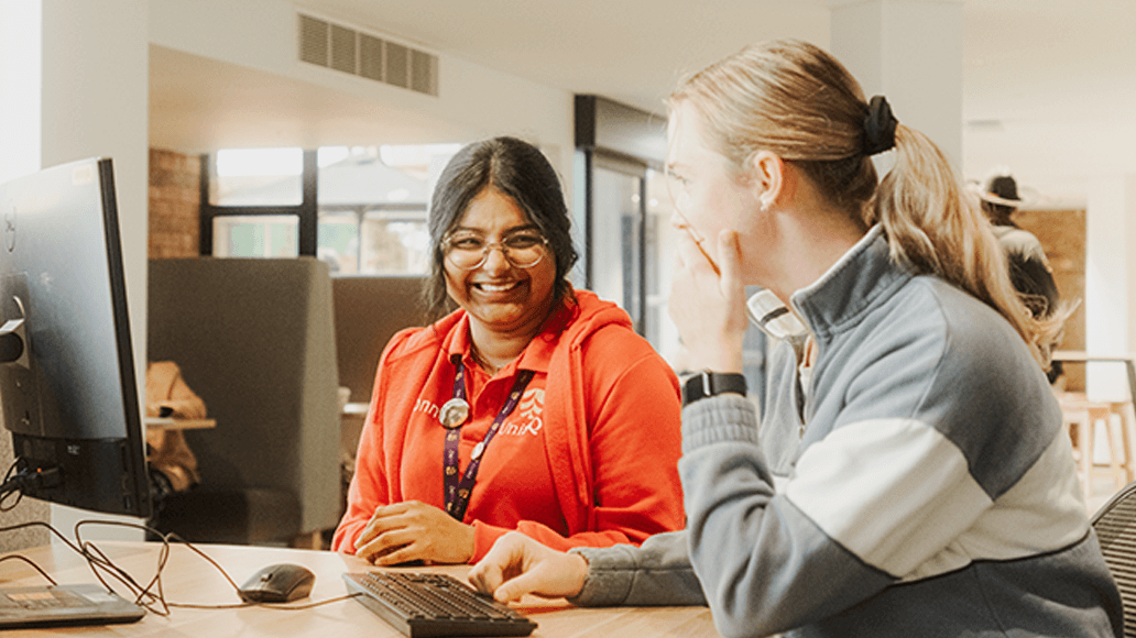 Two people sit at a desk in front of a computer, smiling and conversing in a brightly lit indoor setting.