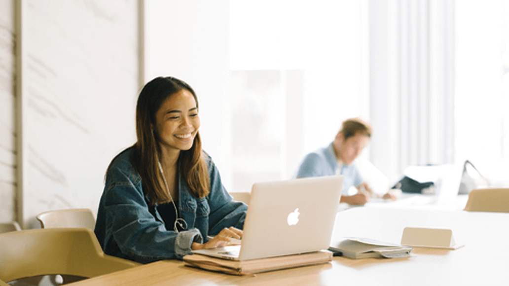 A woman sits at a table using a laptop, smiling, with books and headphones nearby. Another person is working in the background.