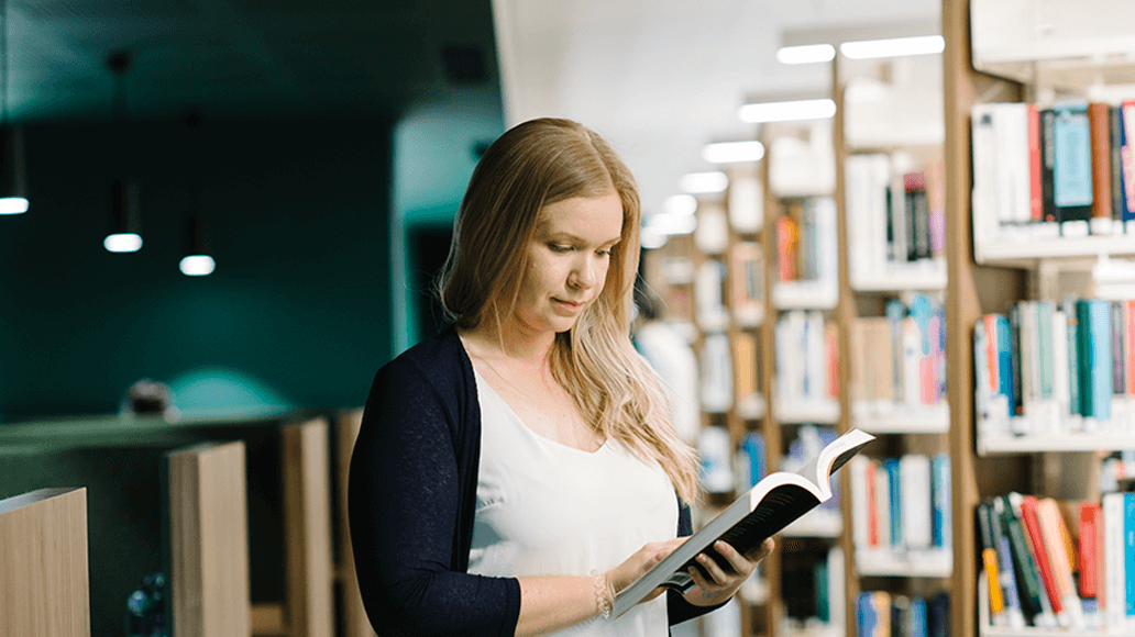 A woman with long blonde hair stands in a library aisle, reading an open book, with shelves of books visible in the background.