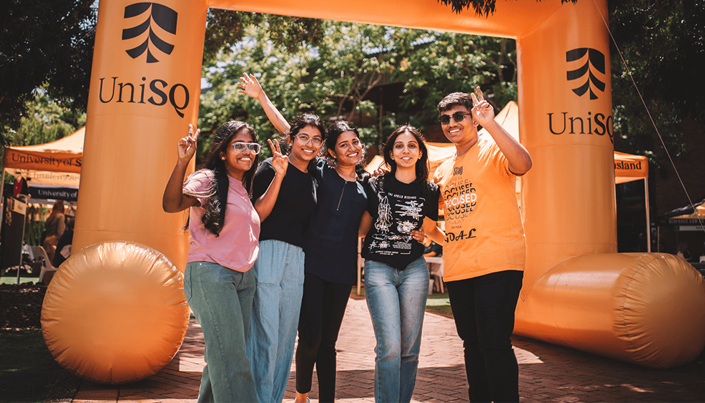 Five people stand together smiling under a large orange inflatable arch with "UniSQ" branding at an outdoor event.