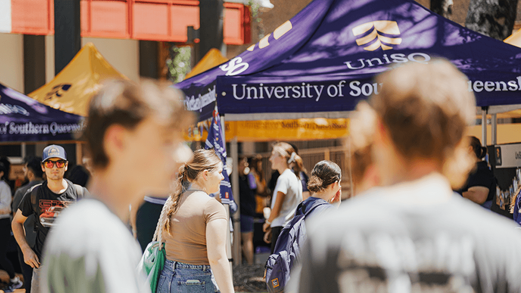 Students walk among university tents, including one with the University of Southern Queensland logo, at an outdoor campus event.