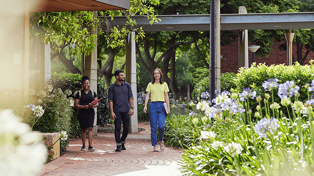 Three people walk along a brick pathway surrounded by greenery and flowers on a sunny day at UniSQ Toowoomba campus.