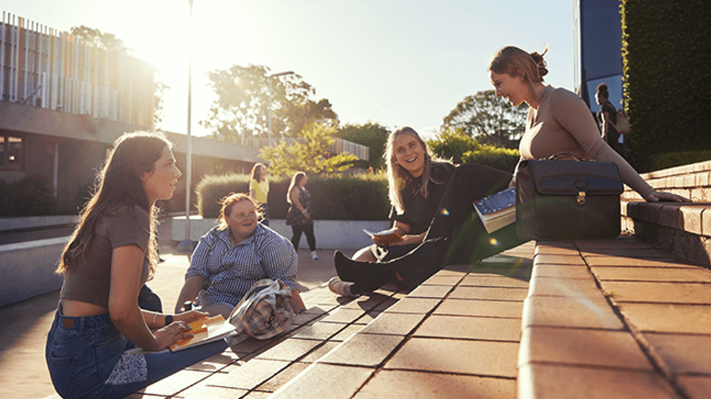 Four people sit and talk together outdoors on steps in sunlight, with books and a bag nearby, next to a modern building.