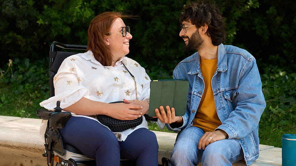 A woman in a wheelchair and a man with a tablet sit on a bench outdoors, smiling and talking to each other.