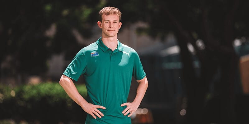 A man stands outdoors with his hands on his hips, wearing a green Puma polo shirt and looking at the camera. Trees and blurred greenery are in the background.