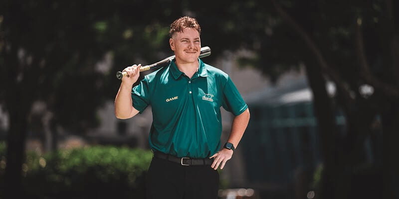 A person in a green polo shirt stands outdoors holding a baseball bat over their shoulder, with one hand on their hip.